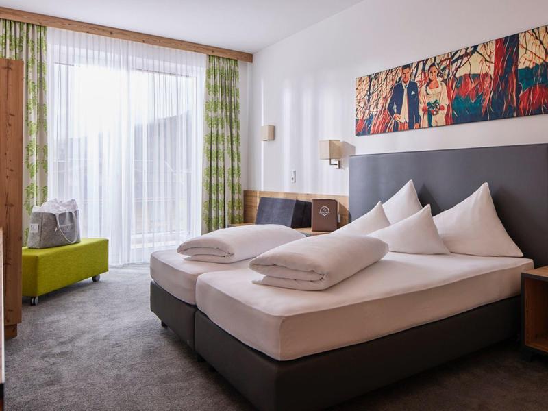 Modernes Schlafzimmer mit Doppelbett, grauem Teppich, grünem Sessel und farbenfrohem Wandbild.