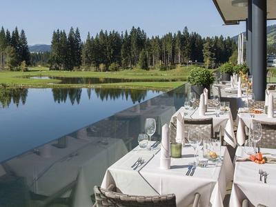 Terrasse mit gedeckten Tischen neben einem ruhigen See vor bewaldeten Hügeln.