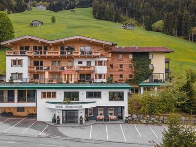 Großes Chalet mit Holz-Balkonen vor grünen Hügeln und Wald im Hintergrund, leerer Parkplatz im Vordergrund.