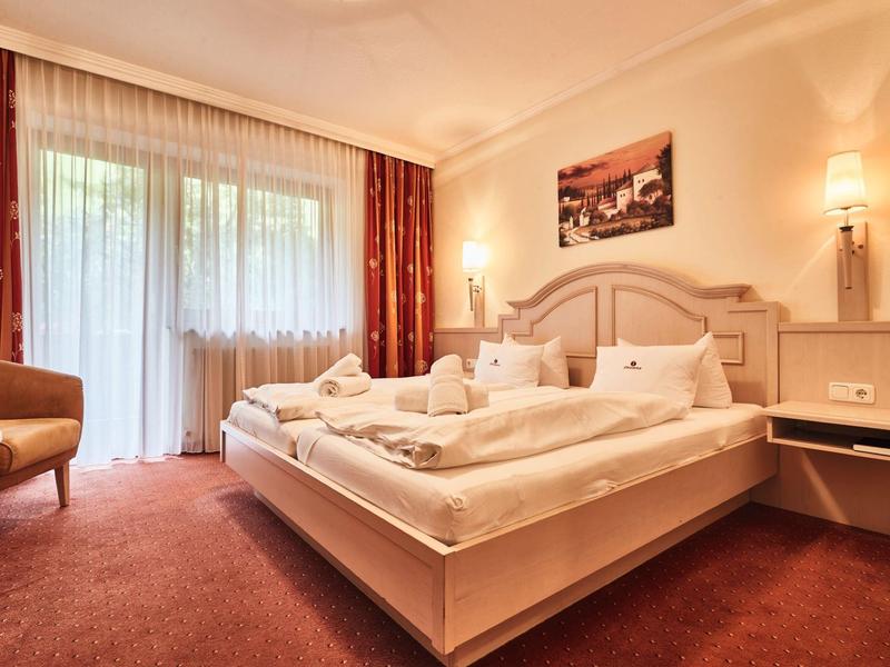 Bright hotel room with double bed, bedside tables, chairs, and large windows with curtains.