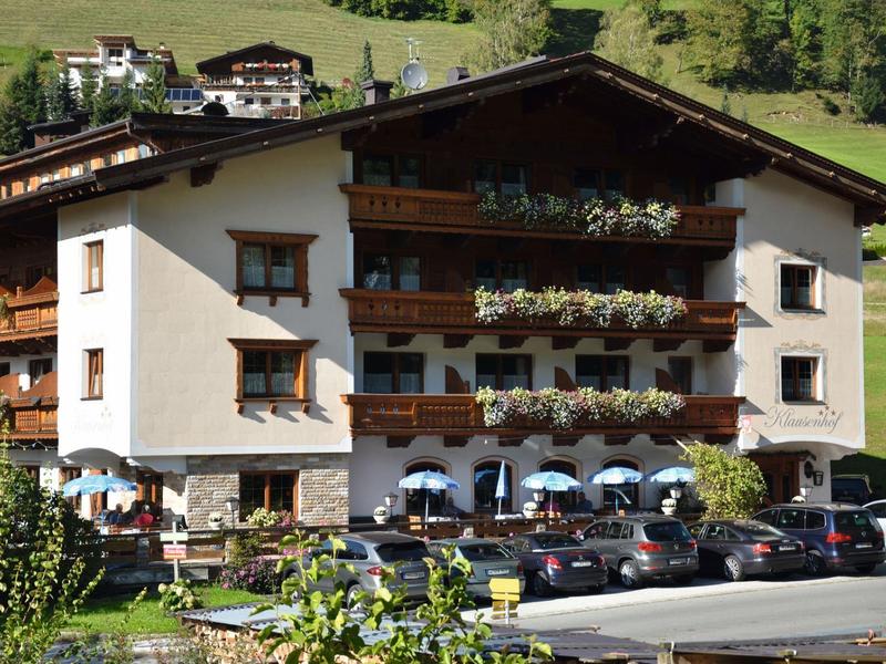 Mehrstöckiges Haus mit Blumen an Balkonen, Autos davor, grüner Hügel im Hintergrund.