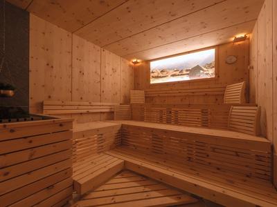 Helle Holzsauna mit mehrstufigen Sitzbänken und beleuchtetem Panoramafenster zeigt Berglandschaft.