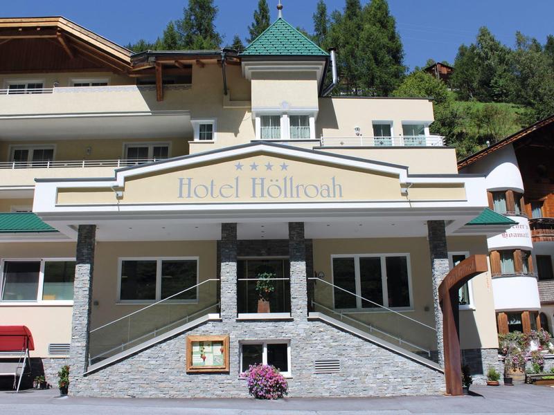 Hellgelbes Hotelgebäude mit grünem Dach und 3 Etagen vor bewaldetem Hügel unter blauem Himmel.