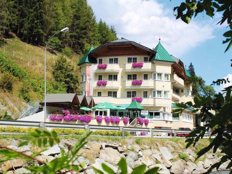Mehrstöckiges Hotel mit Balkonblumen vor bewaldetem Hügel und Steinmauer im Vordergrund.