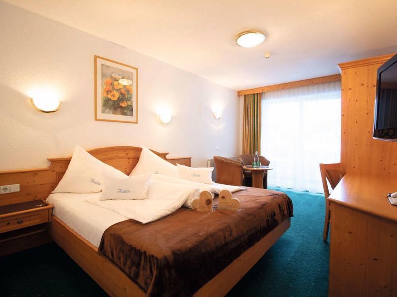 Gemütliches Hotelzimmer mit Doppelbett, brauner Bettdecke, Schreibtisch und Balkon vor hellem Vorhang.