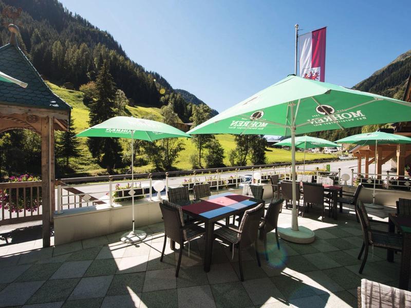 Sonnige Außenterrasse mit Tischen, grünen Sonnenschirmen und Blick auf grüne Hügel und Wald.