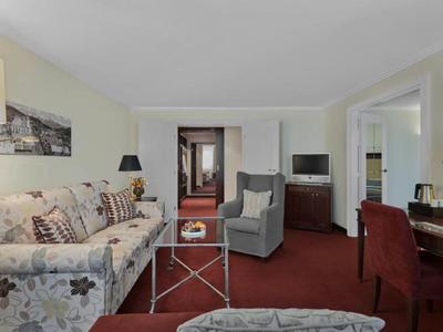 Pohodlný hotelový pokoj s pohovkou, křesly, psacím stolem a televizí na červeném koberci.