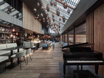 Sala da pranzo moderna in hotel con pannelli in legno, pianoforte a coda e luci a sfera sospese sotto tetto in vetro.