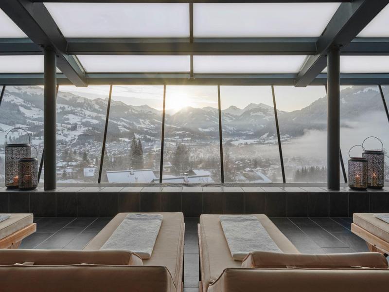 Výhled na zasněžené hory skrz velká skleněná okna z moderního interiéru s pohodlnými lehátky.