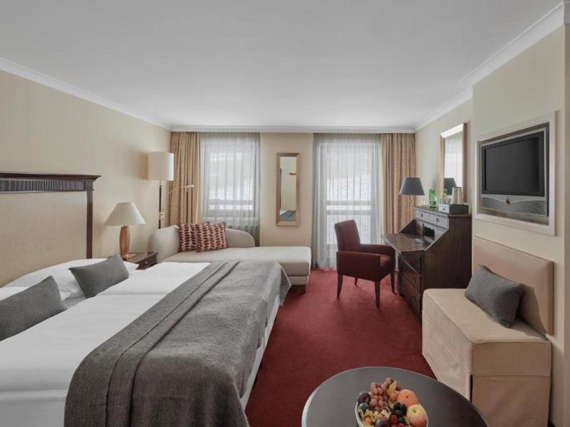 Moderně zařízený hotelový pokoj s manželskou postelí, psacím stolem, televizí a červeným kobercem.