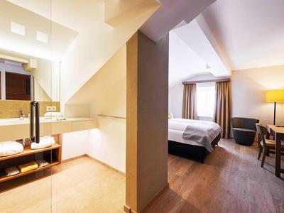 Helles Hotelzimmer mit Bad, Holzfußboden, Doppelbett und Schreibtisch mit Stuhl und Lampe.