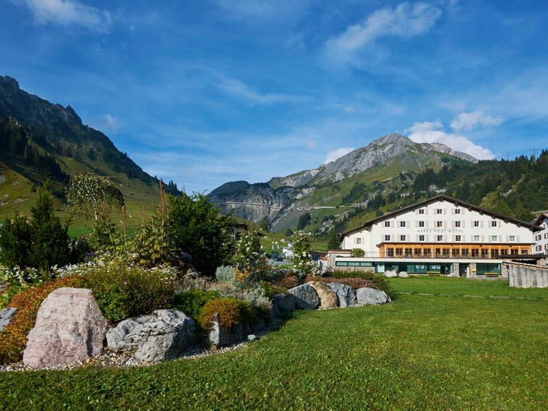 Berglandschaft mit grünem Tal, Felsen, Blumen und großem weißem Hotel unter blauem Himmel.