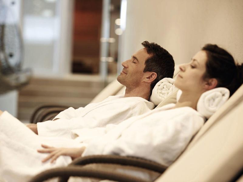 Zwei Personen entspannen in weißen Bademänteln auf Liegestühlen in einem ruhigen Raum.