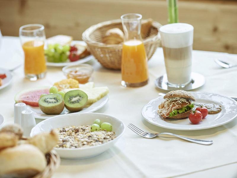 Frühstückstisch mit Orangensaft, Kiwi, Müsli, Brötchen und einem Latte Macchiato auf weißem Tuch.