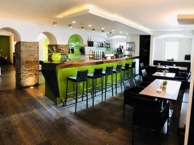 Moderner Barbereich mit grünem Tresen, schwarzen Barhockern und Holzboden in warmem Licht.