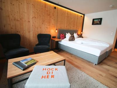 Hotelzimmer mit Holzverkleidung, zwei Sesseln, Bett, Teppich und Hocker mit Aufschrift "HOCK DI HER".