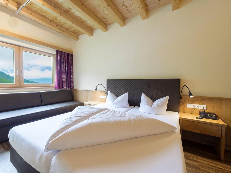 Hotelový pokoj s manželskou postelí, pohovkou, dřevěným stropem a výhledem na hory velkým oknem