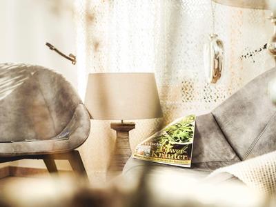 Lichte kamer met twee grijze fauteuils, een lampentafeltje en een boek voor witte gordijnen.