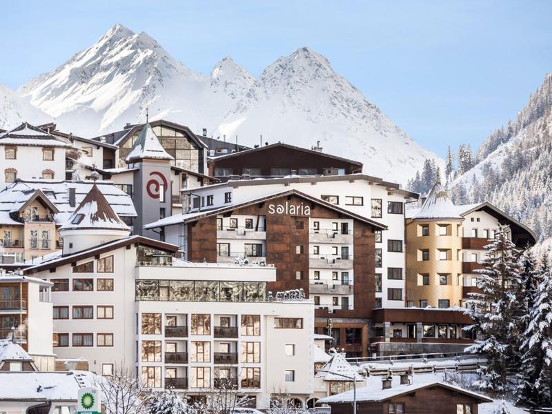 Villaggio montano innevato con chalet davanti a cime innevate sotto un cielo limpido.