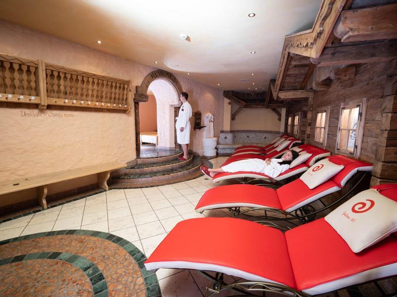 Sala relax con lettini rossi e due persone in accappatoi sullo sfondo.