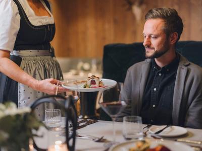 Ein Mann sitzt am gedeckten Tisch, eine Kellnerin serviert ihm ein Dessert auf weißem Teller.