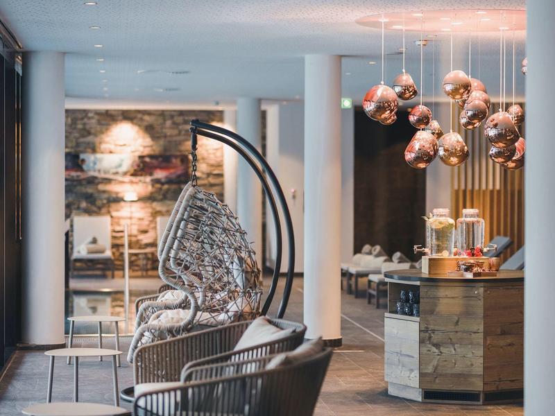 Moderner Innenraum mit Hängekorb, gemustertem Sitzkissen und kupferfarbenen Hängelampen am Holztresen.