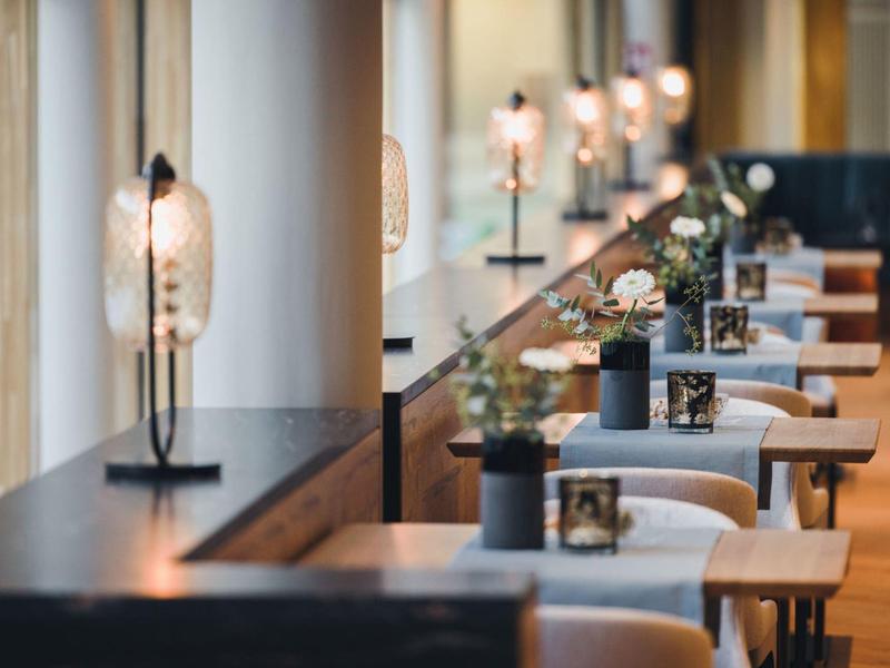 Moderner Gastraum mit langen Fenstern, Tischen, Stühlen und dekorativen Lampen in warmem Licht.