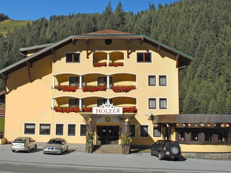 Bâtiment d'hôtel jaune devant une colline boisée avec plusieurs voitures garées.