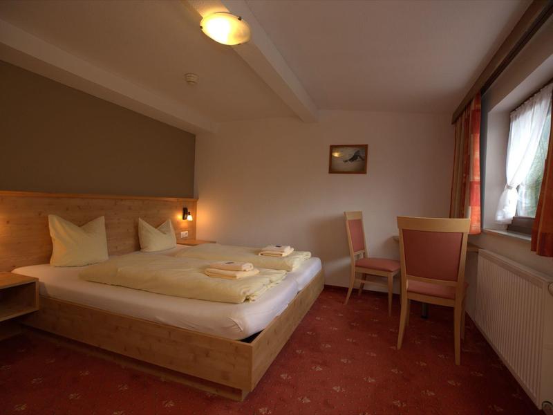 Einfaches Hotelzimmer mit Doppelbett, roten Teppichboden, zwei Stühlen und einem Fenster mit Vorhängen.