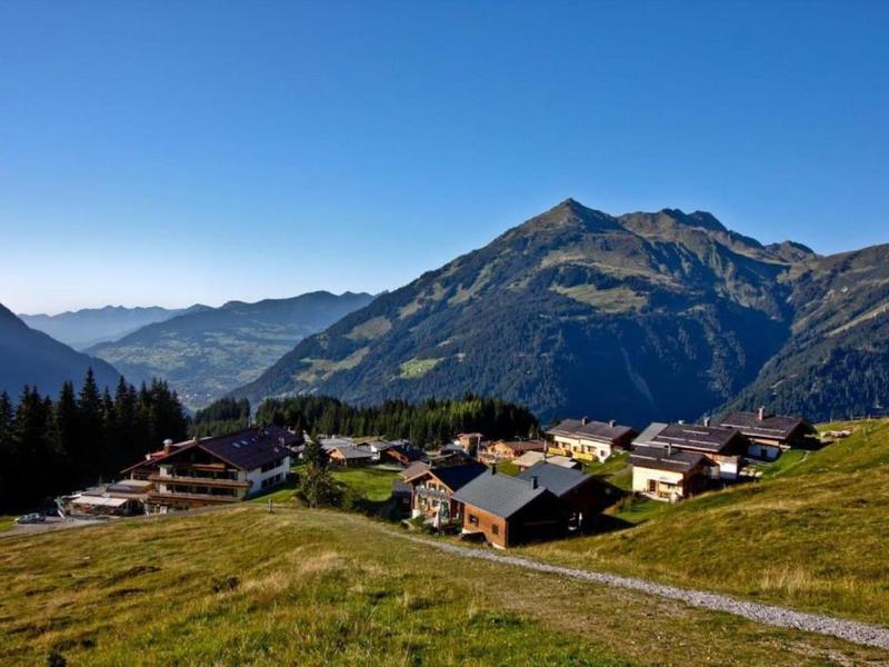 Landschaft mit grünen Hügeln, kleinen Häusern und hohen Bergen unter klarem blauem Himmel.