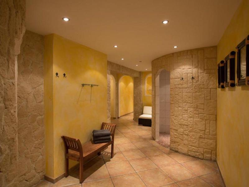 Flur mit gelben Wänden, Rundbögen, Steinstruktur und Holzbänkchen mit Kissen. Deckenlampen leuchten.