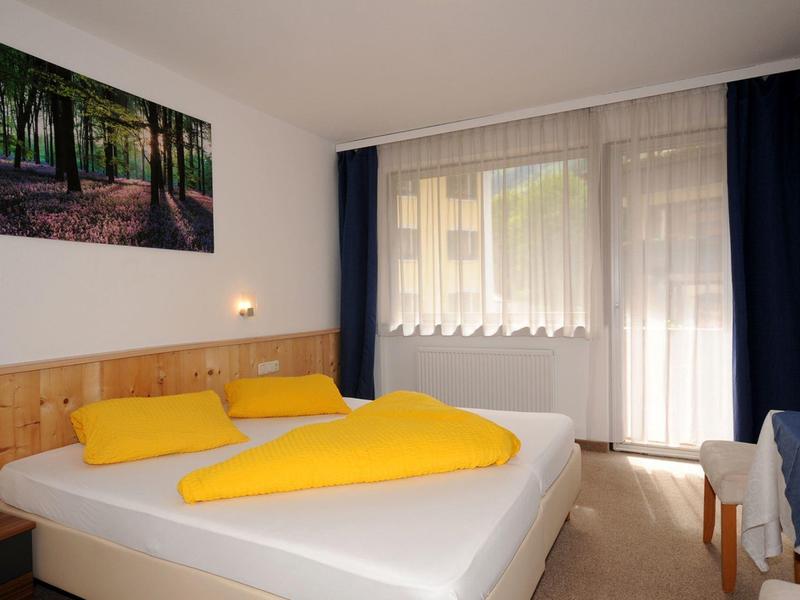 Hell eingerichtetes Schlafzimmer mit Doppelbett, gelber Bettwäsche und großem Fenster mit Vorhängen.