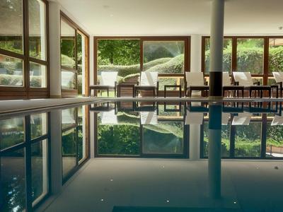 Innenraum eines Schwimmbads mit klarer Wasseroberfläche, großen Fenstern und Holzstühlen am Rand.