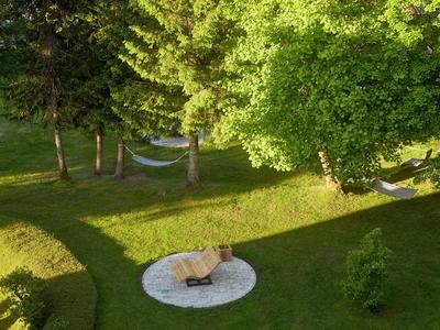 Jardin vert avec arbres, pelouse et deux chaises sur une terrasse blanche ronde au soleil.