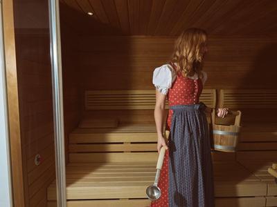 Frau in traditionellem Kleid steht mit Holzeimer und Kelle in einer Sauna mit Holzbänken.