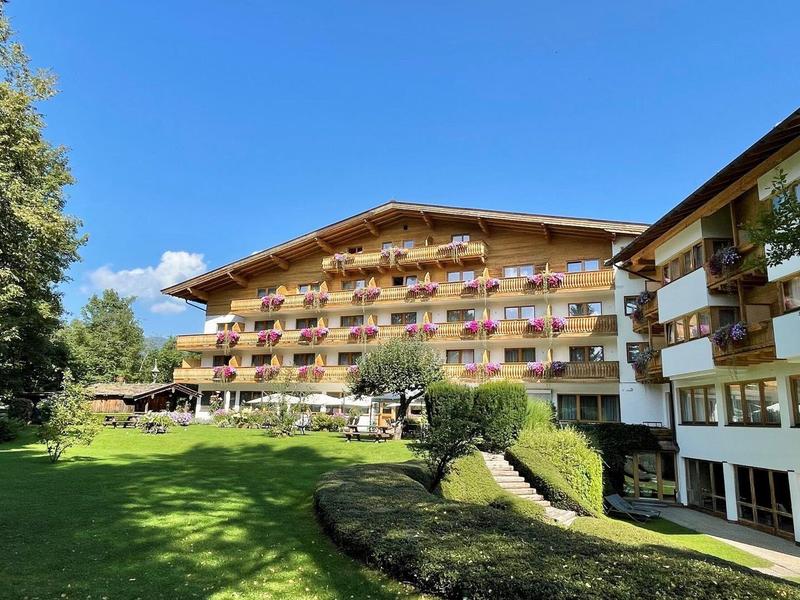 Großes Alpenhotel mit Balkonen, Grillplatz auf dem grünen Rasen und klarem blauem Himmel.