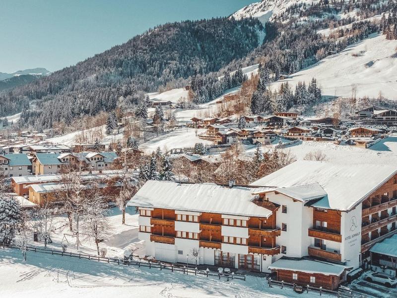 Winterliches Dorf mit schneebedeckten Bergen, Häusern und kahlen Bäumen im sonnigen Tal.