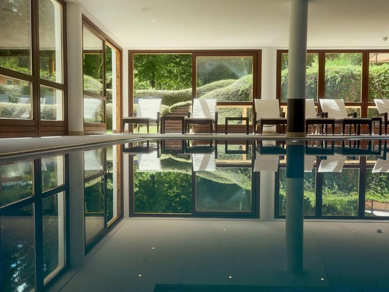 Innenraum eines Schwimmbads mit klarer Wasseroberfläche, großen Fenstern und Holzstühlen am Rand.