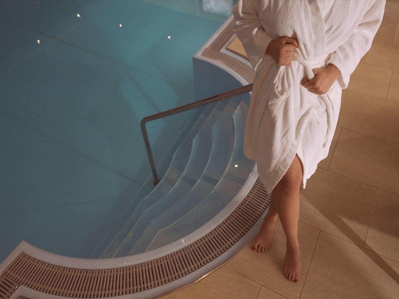 Person in weißem Bademantel steht barfuß neben beleuchtetem Schwimmbecken mit Wasser.