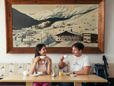 Zwei Personen sitzen an einem Tisch und trinken Kaffee vor einem großen Berggemälde.