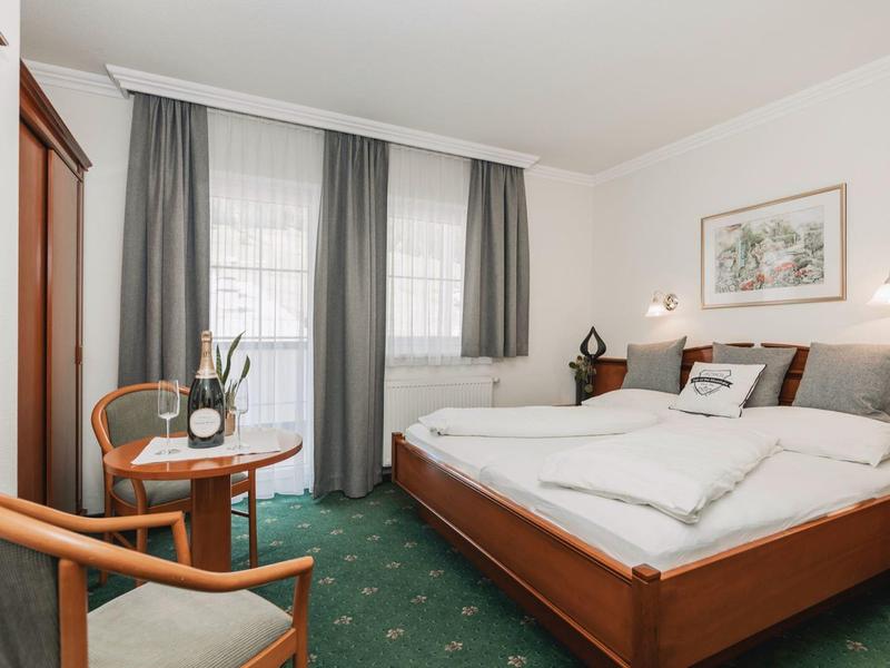 Gemütliches Hotelzimmer mit Doppelbett, Holzmöbeln, grünem Teppich und Fenster mit Vorhängen.