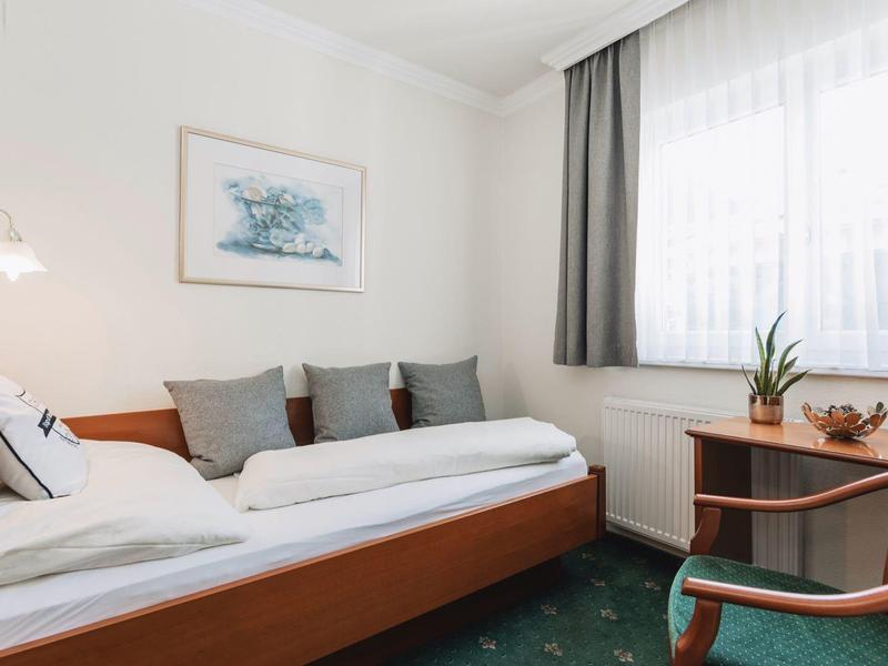 Kleines Zimmer mit Einzelbett, grünem Teppich, Holzmöbeln und Fenster mit Gardinen.