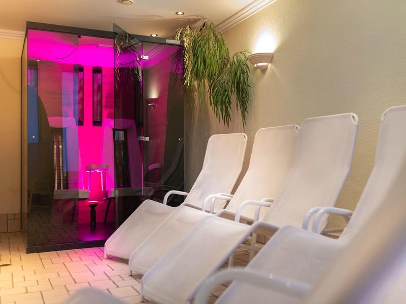 Ruheraum mit weißen Liegestühlen und einer beleuchteten Infrarotsauna in warmem, gedämpftem Licht.