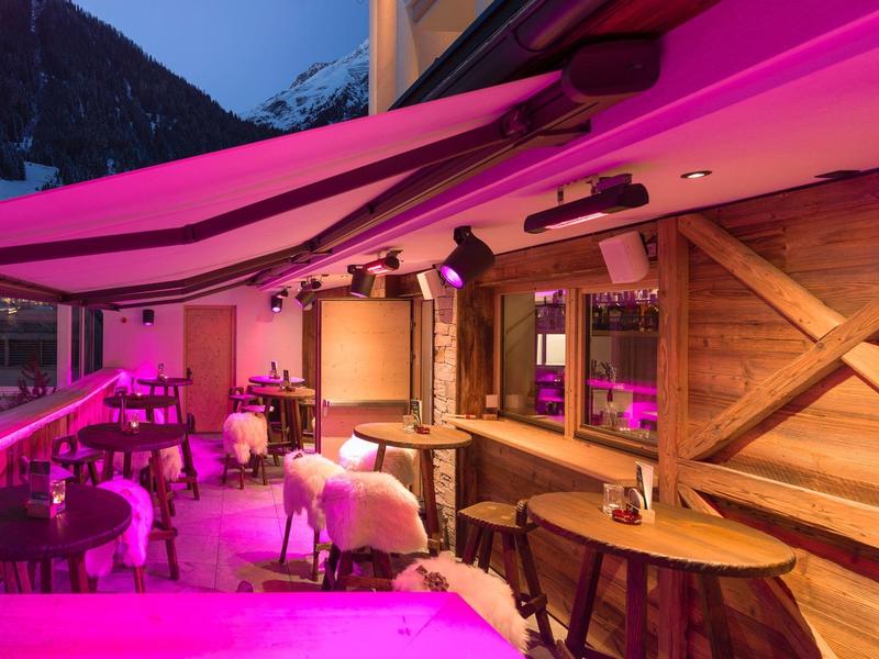 Gemütliche, holzverkleidete Terrasse mit Tischen, Stühlen und rosa Beleuchtung am Abend.