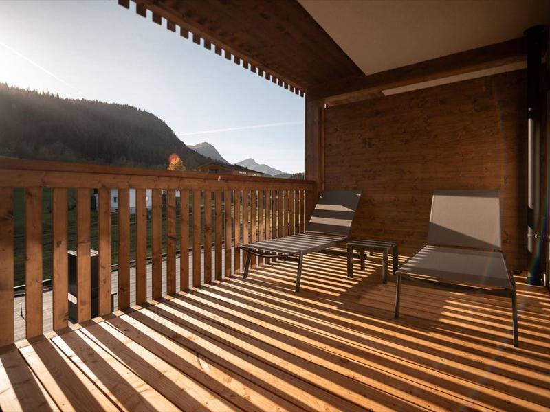 Sunlit balcony with two lounge chairs and mountain view under wooden railing shadows.
