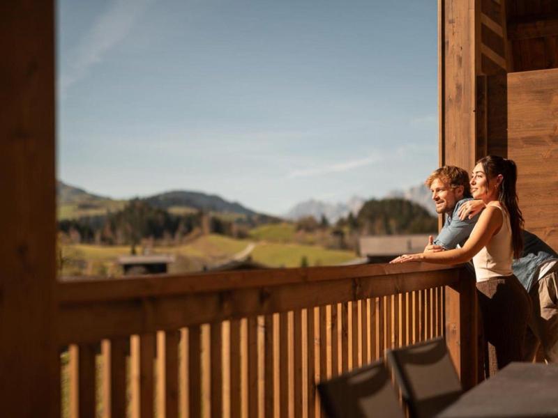 Two people stand on a wooden balcony overlooking a scenic rural landscape on a sunny day.