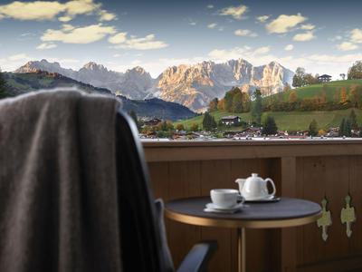 Balcón con mesa, tetera y taza, vista a colinas verdes y montañas iluminadas por el sol.