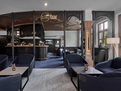 Bar elegante de hotel con sillones azules, muebles de madera y grandes ventanas que permiten luz natural.