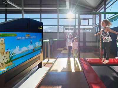 Niños juegan mini golf en un espacio interior luminoso con grandes ventanas y sol.