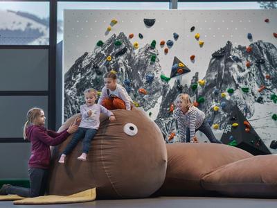 Niños y adultos escalando una estructura grande marrón en un gimnasio de escalada con fondo de montañas.